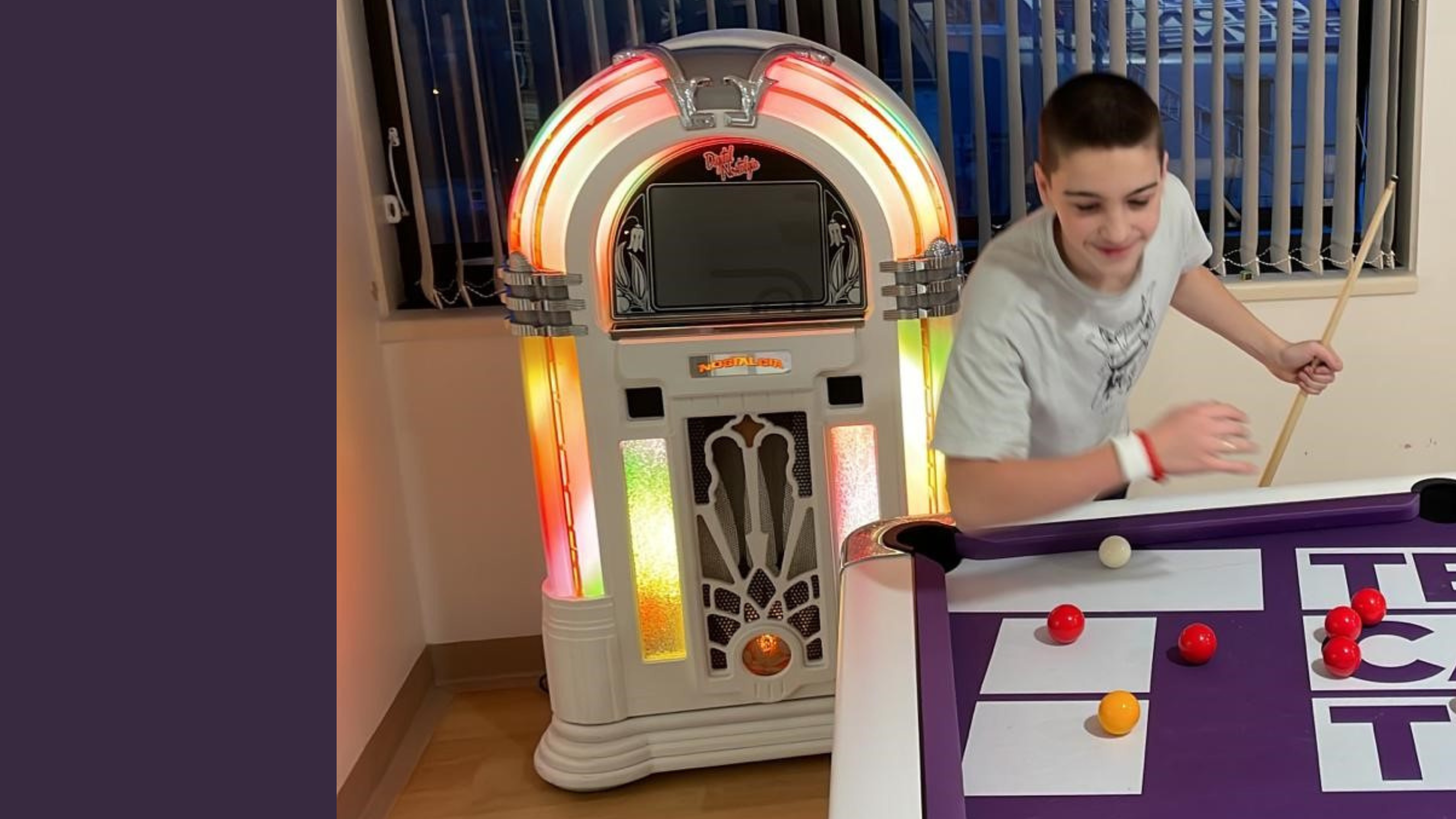 A boy playing pool on a purple pool table that says Teenage Cancer Trust. Behind him there is a brightly lit juke box