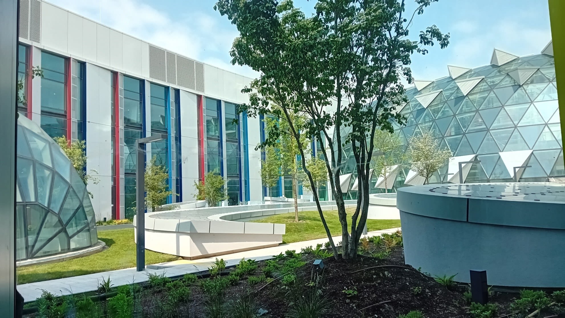 A single storey brightly coloured building with lots of glass, next to a glass dome that looks a bit like the Eden Project in Cornwall. There is a landscaped garden with trees and simple foliage