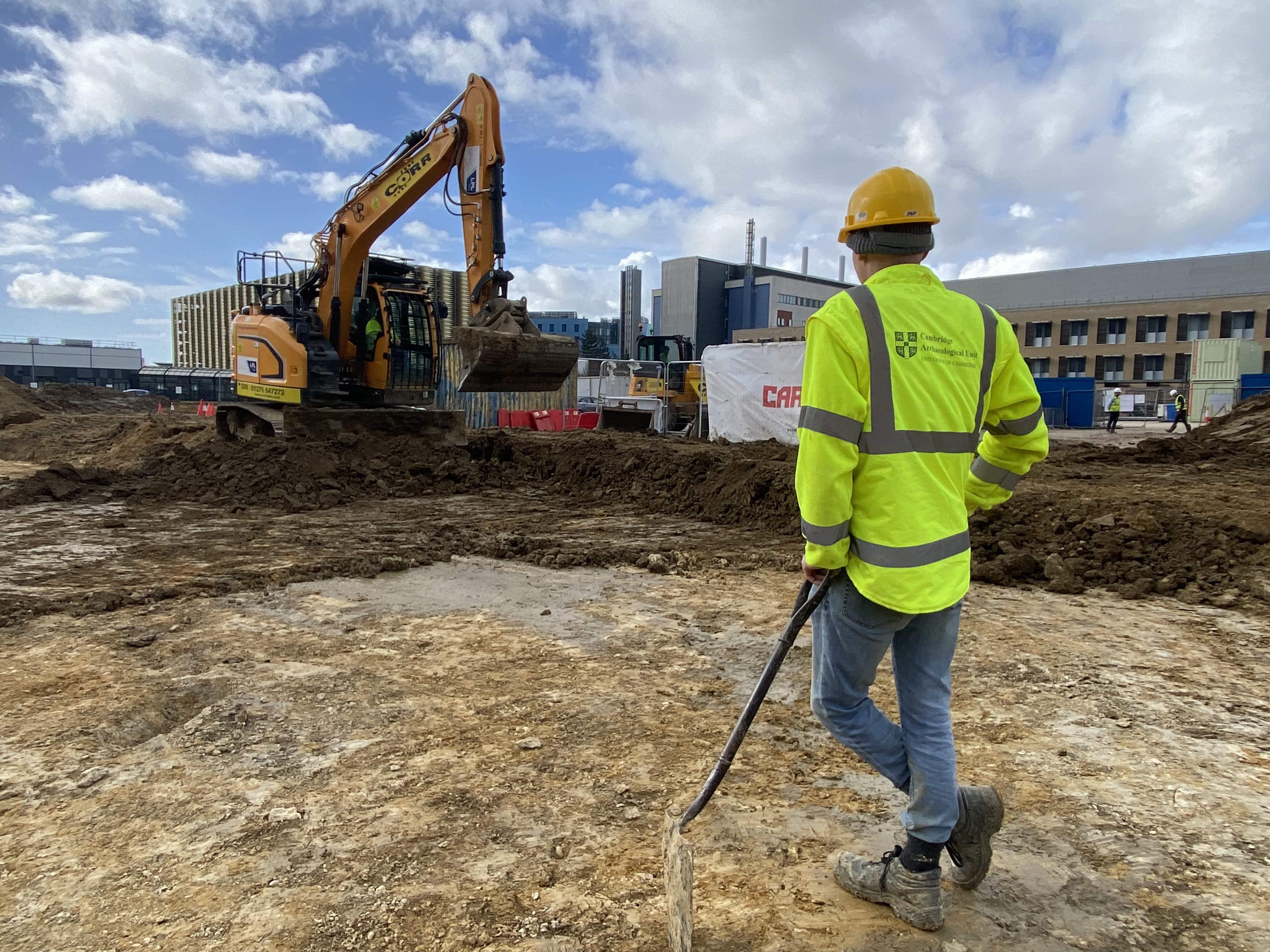 An archaeologist in a reflective jacket, hard hat and jeans stands with a shovel looking at a huge digger excavating the mud on the Cambridge Children's Hospital site.