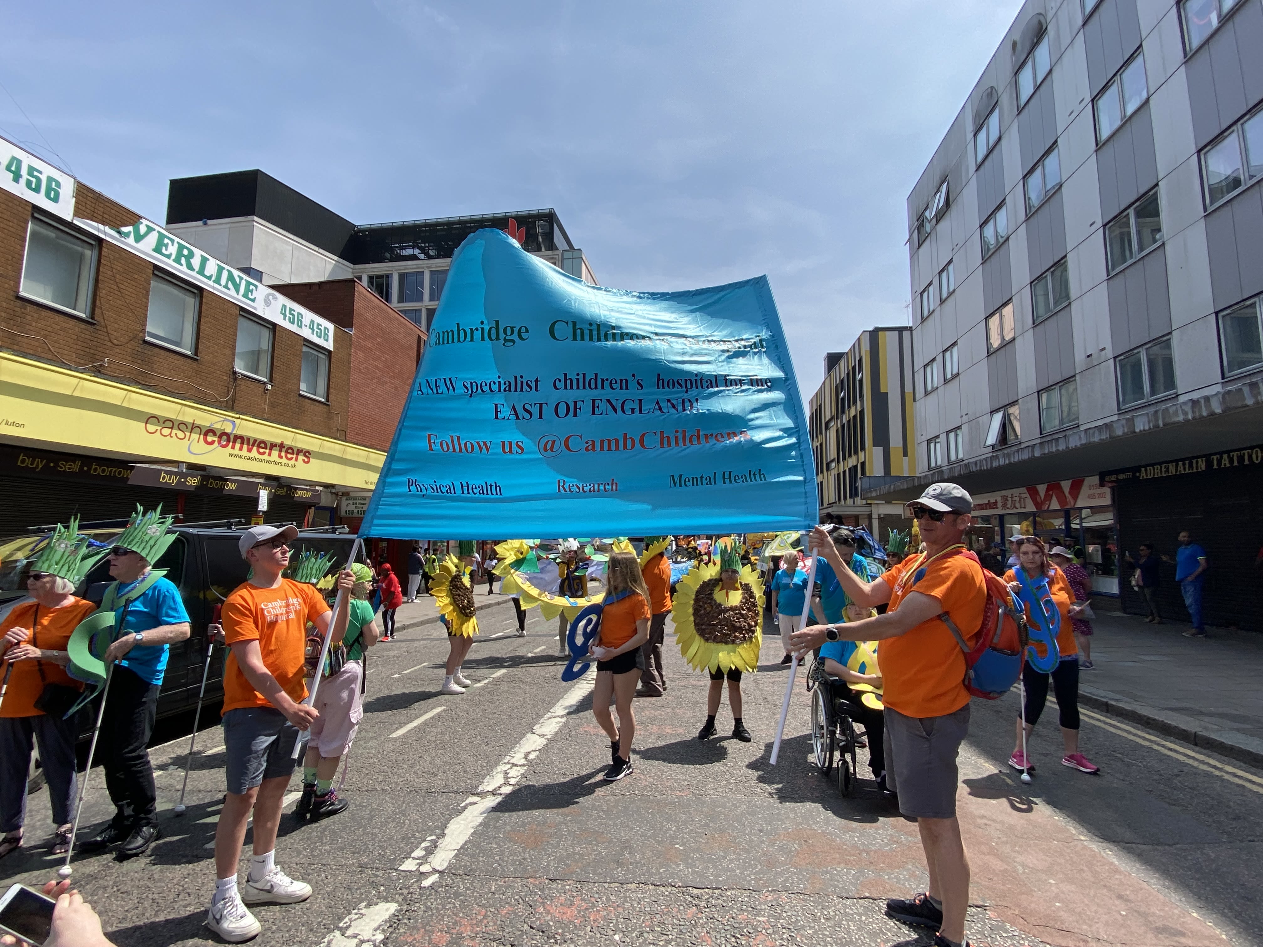 Two people holding up a large blue Cambridge Children's Hospital banner. They are standing on a street in the sunshine waiting for the carnival parade to start