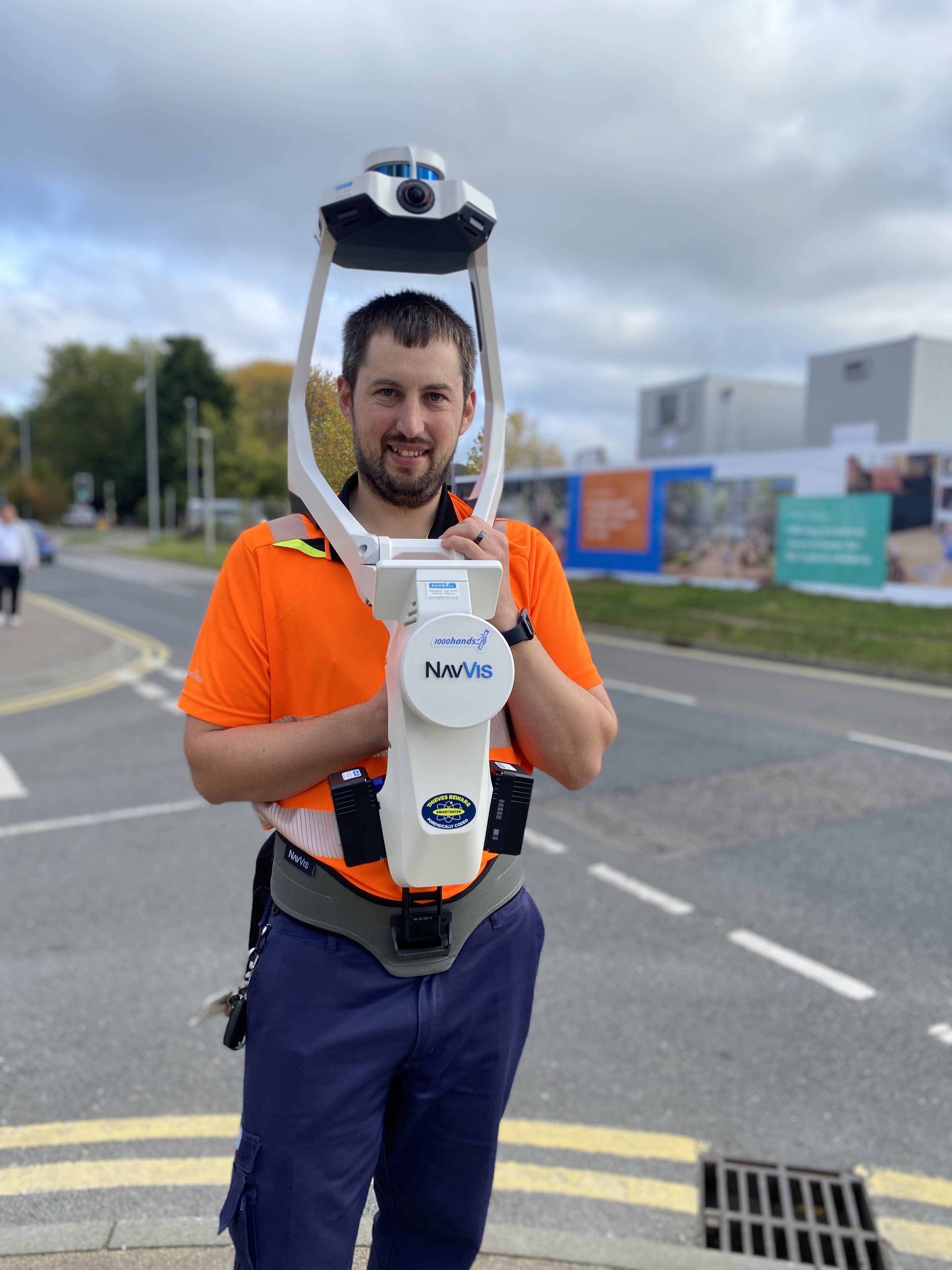 A man with dark hair and beard, and orange t shirt, standing next to a building site area. He is wearing specialist surveying equipment which is like a big white robot machine on his front. There are two poles each side of his head with a camera on top, that looks a bit space age.