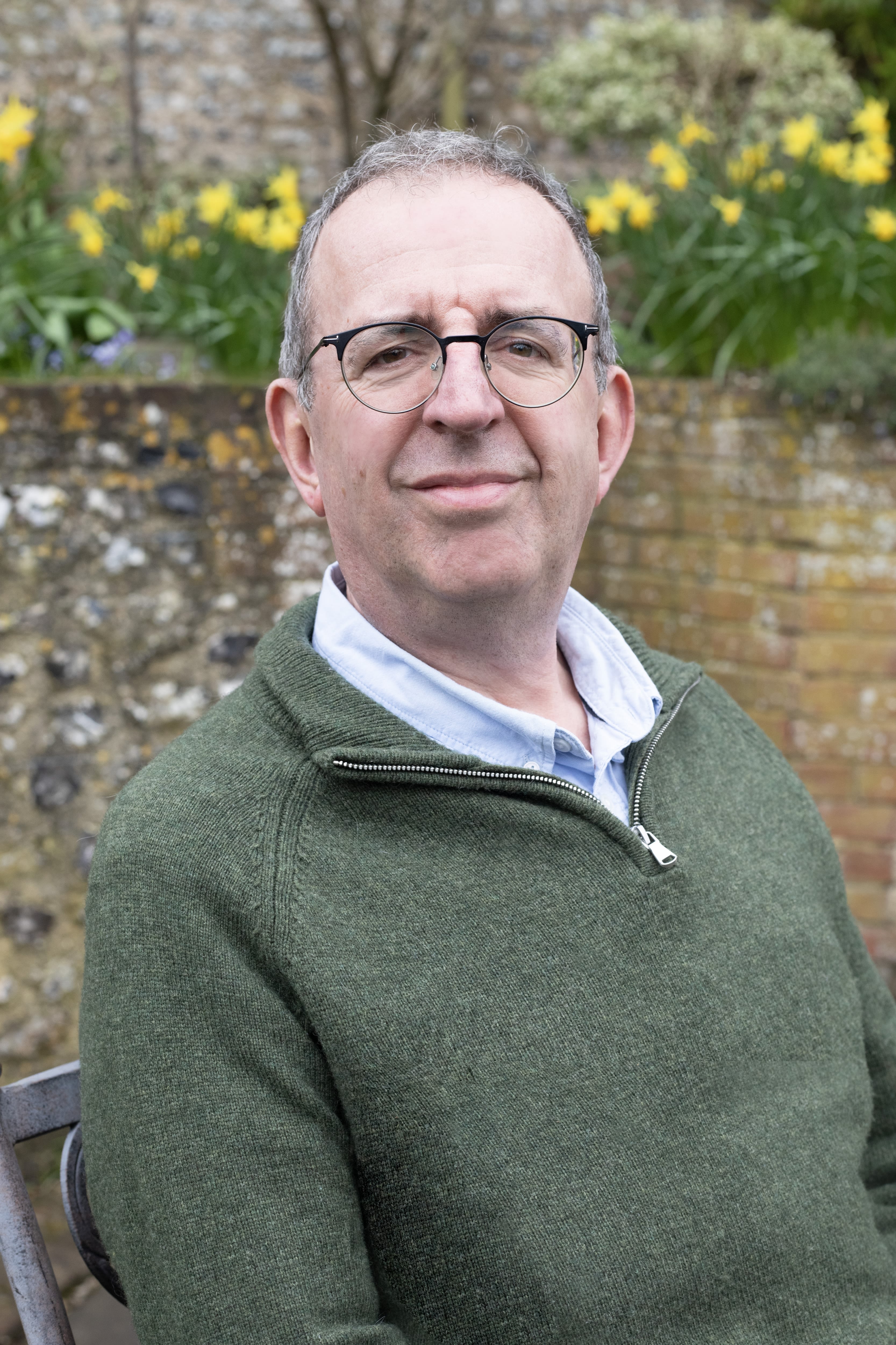 A man with grey receding hair and glasses standing in front of a red brick wall with daffodils tumbling over the top. He is wearing a moss green jumper and pale blue shirt