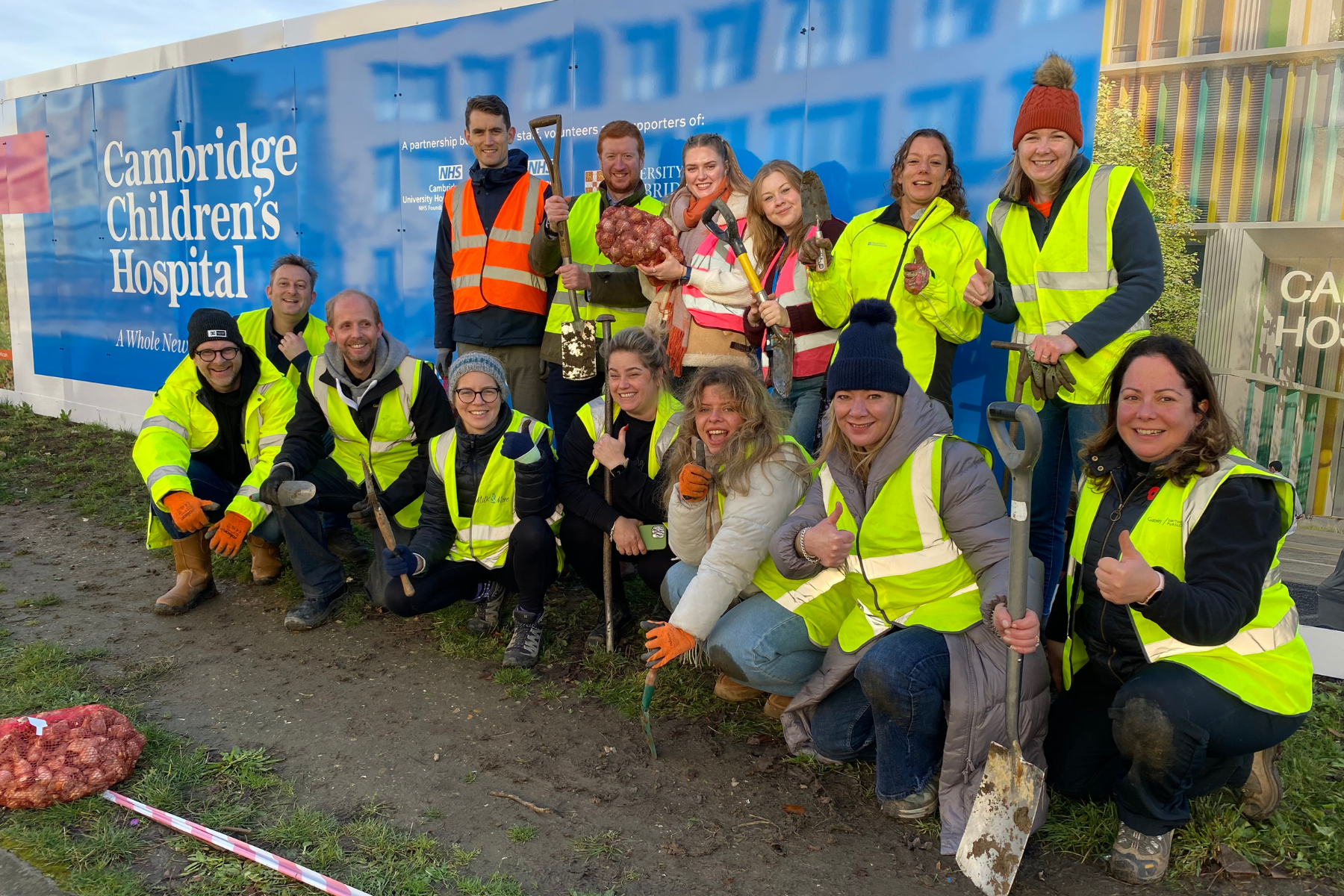 A group of people in high vis jackets and warm clothes holding spades and trowels and bulbs in front of the Cambridge Children's Hospital hoardings