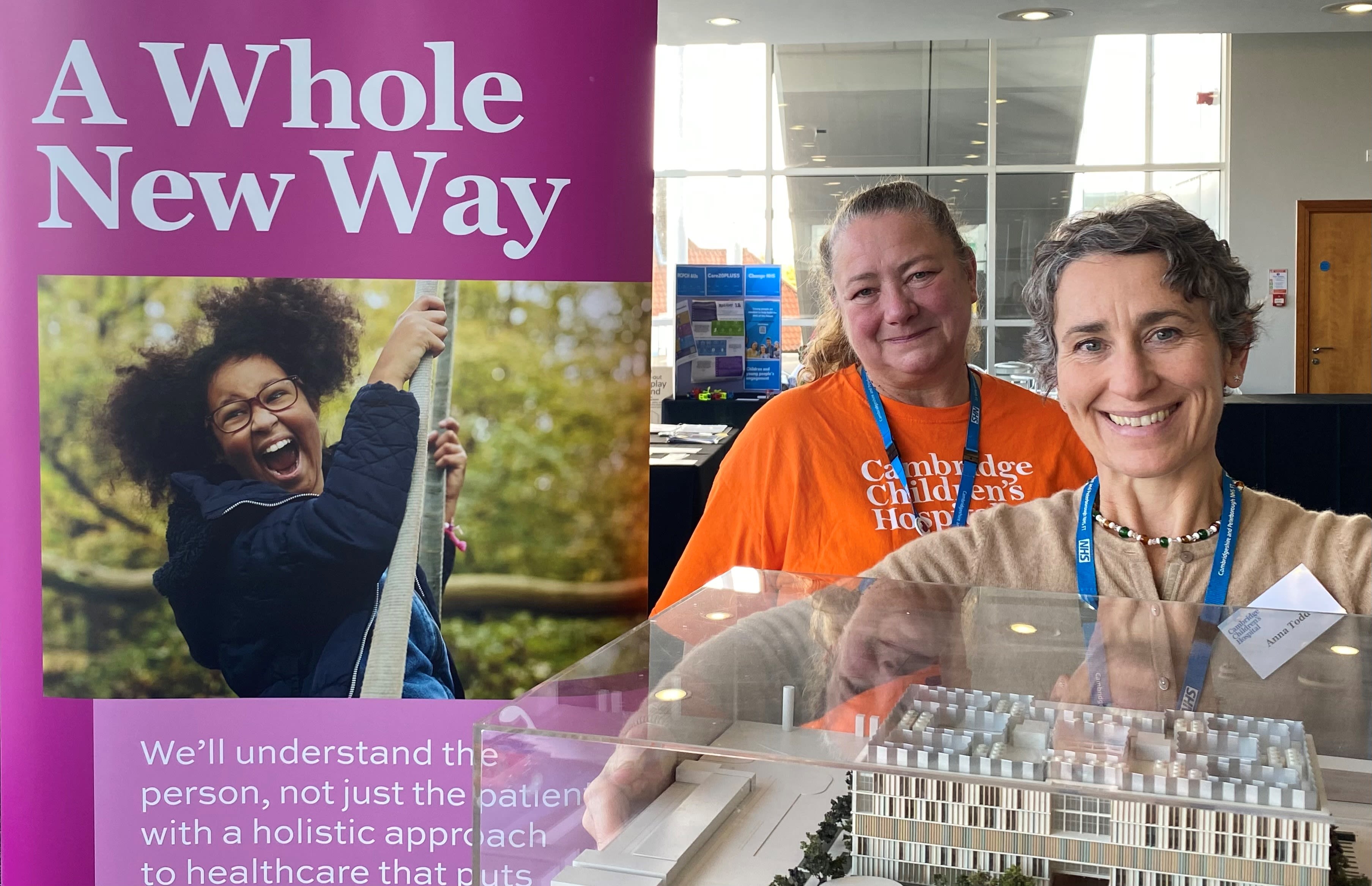 Two middle aged women smiling at the camera, with a pink banner behind them that has a photo of a young girl climbing a rope. It says A Whole New Way. One woman is holding a model of Cambridge Children's Hospital in a perspex box