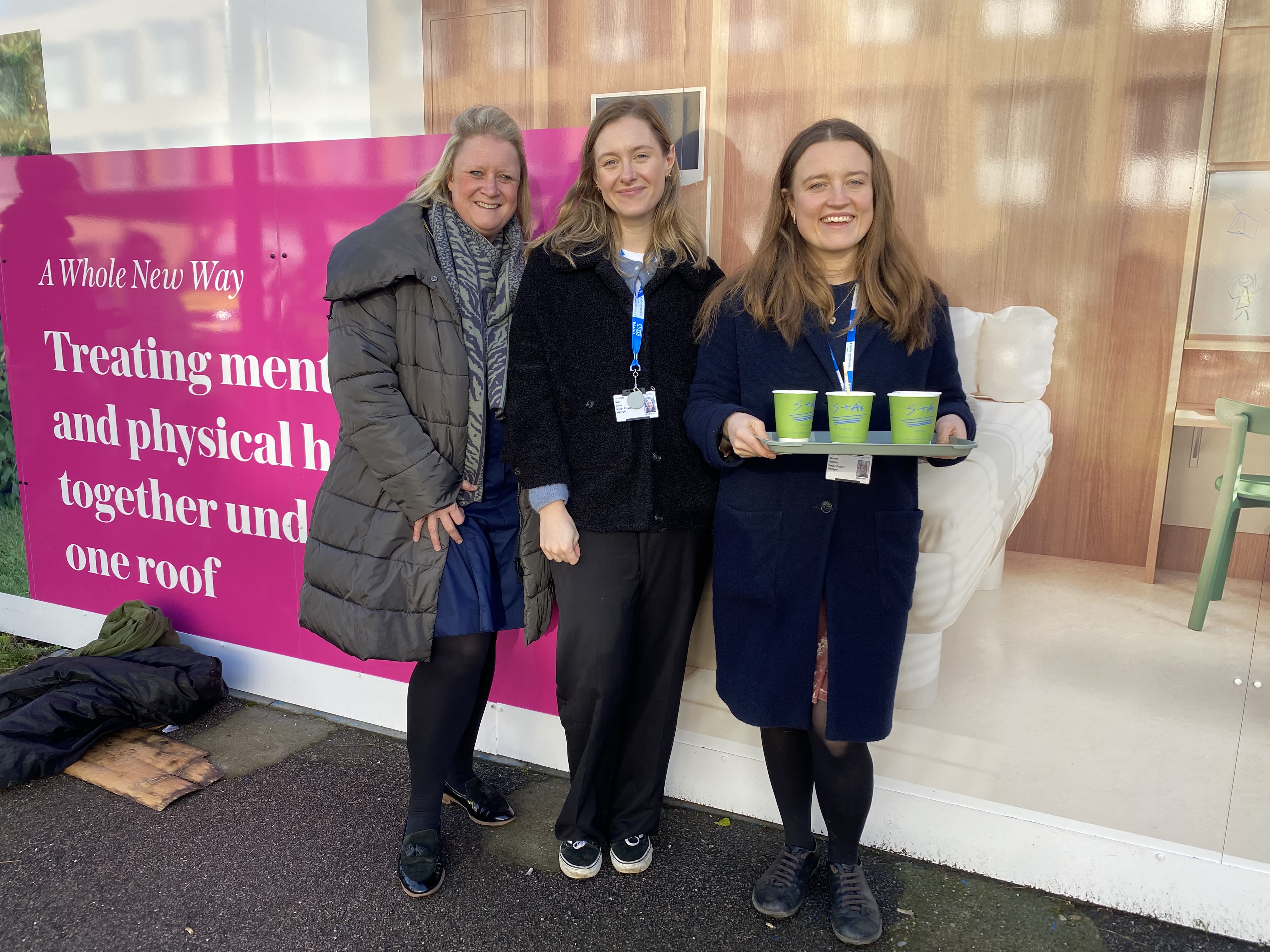 Three women smiling at the camera at an outside event. One of them is holding a tray of tea cups