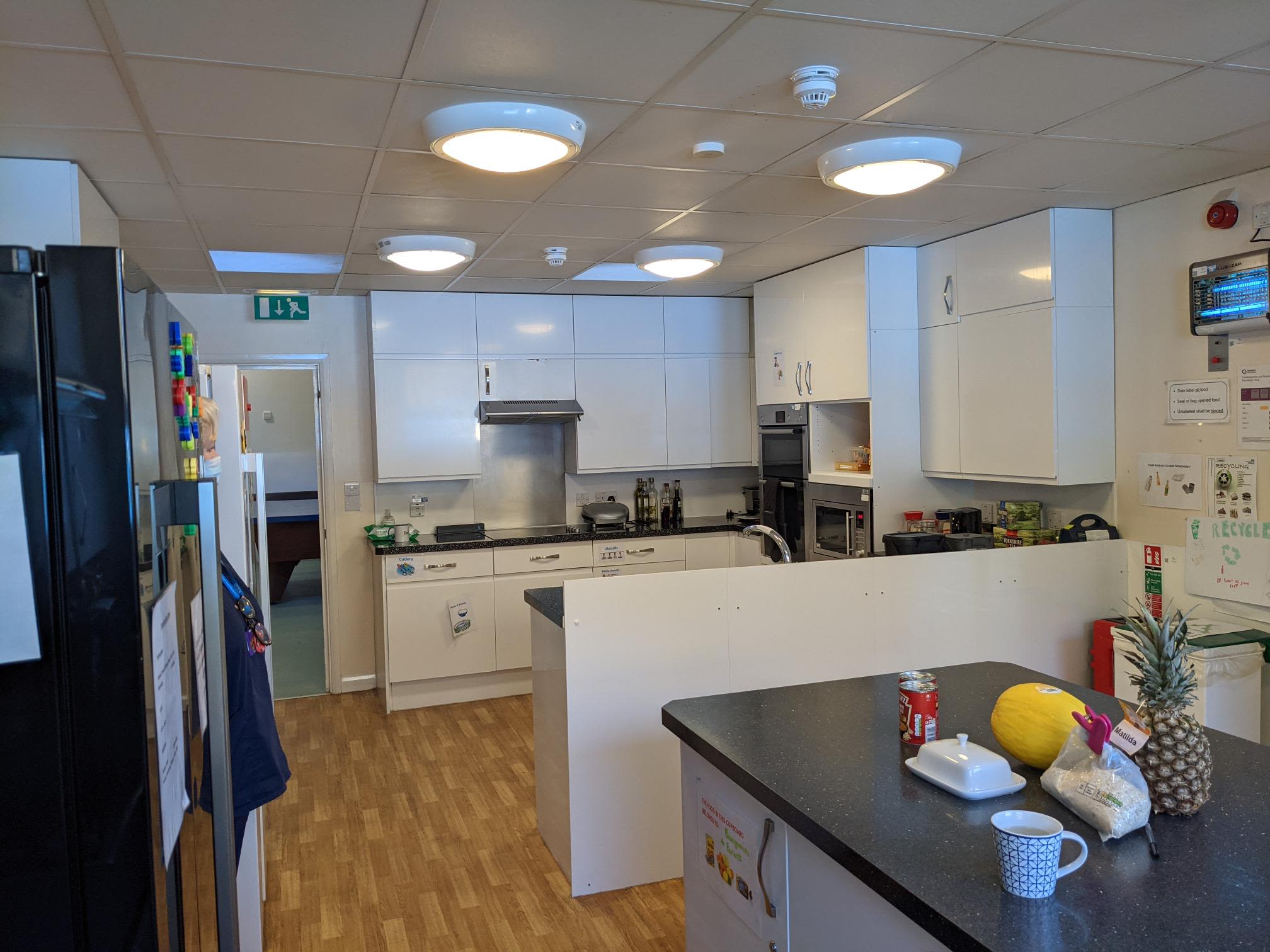 A modern kitchen in a mental health ward. There are white kitchen cabinets and black work surfaces. There is fruit and drinks on the kitchen island, including a pineapple and a melon