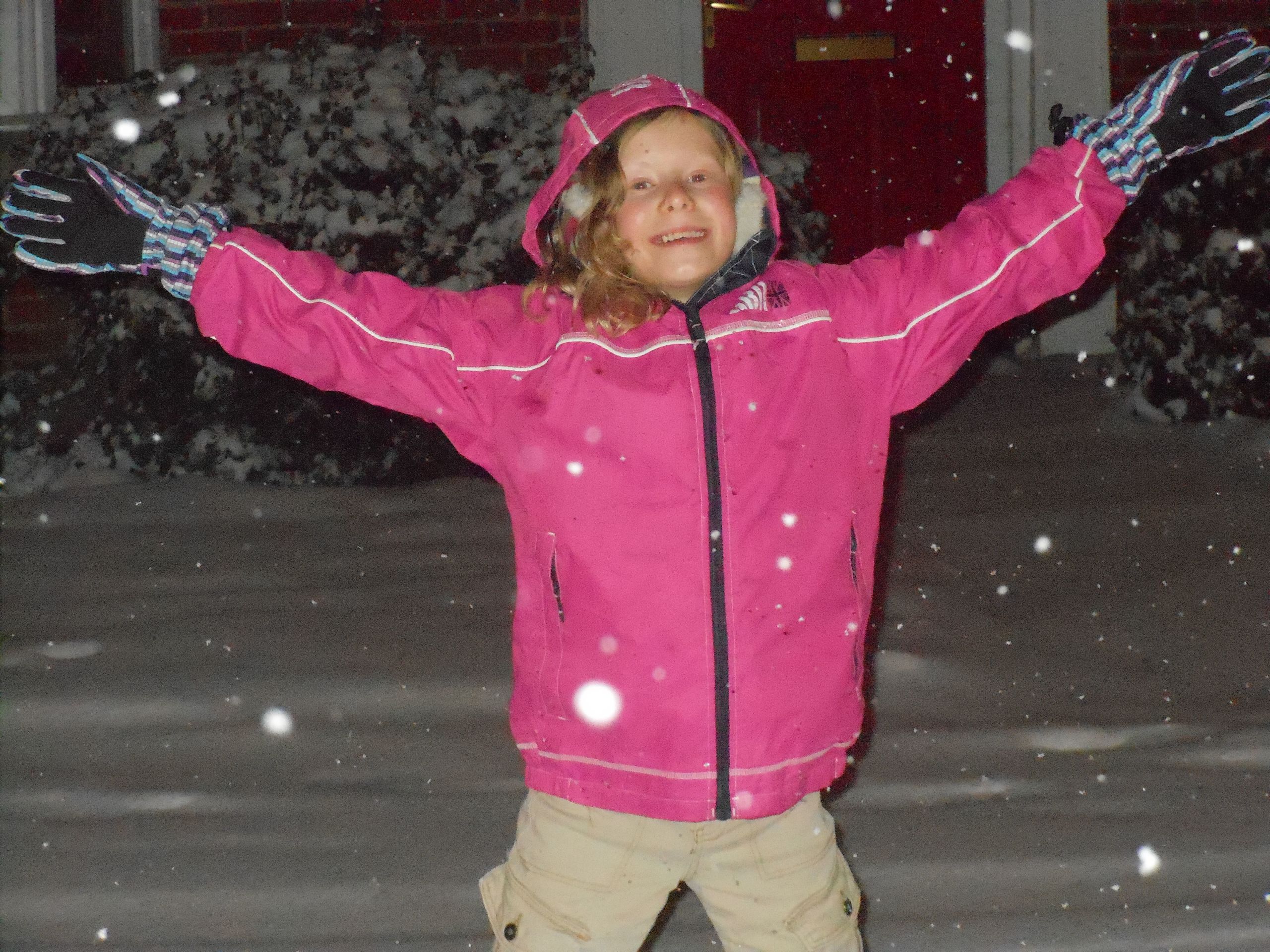 A young jess with a beaming smile standing in the snow, arms extended. Her hair is long and curly