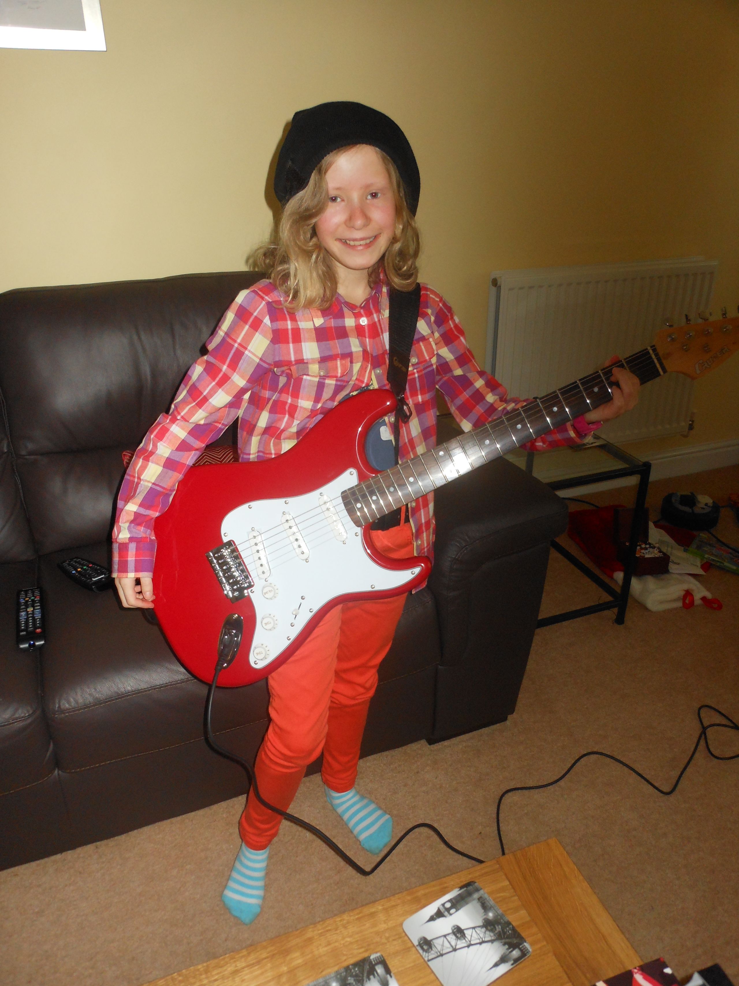 A young jess, with a big smile, holding her red electric guitar. She is wearing a black beanie hat and a red checked shirt.