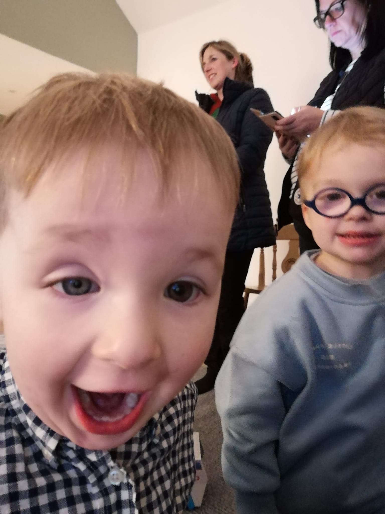 Two very young boys. One has fair straight hair and is wearing a black and white checked shirt. His face is close to the camera and he's doing a big open mouthed smile. The other little boy has on black framed oval glasses and a grey sweatshirt. He is a little further away from the camera.