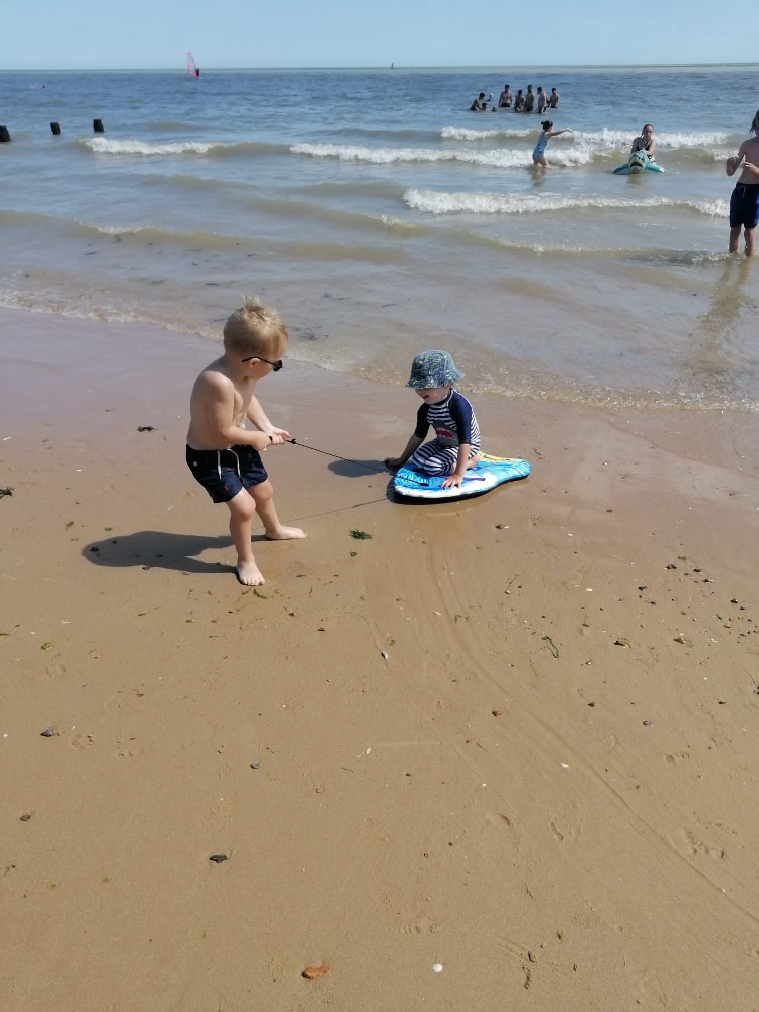 An English beach scene with two little boys playing on the sand. One boy is wearing black trunks and goggles. He is pulling another boy along on a body board. The other little boy has on a blue sunhat and an all in one navy swimsuit. It is a very sunny day and the sea has gentle waves, with people swimming