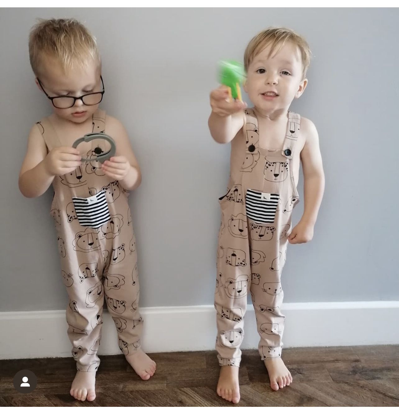 Two little boys in matching animal print dungarees. One boy has black rimmed glasses and is looking at a toy in his hand. The other boy is looking at the camera and handing a green toy to the photographer. They are standing against a pale grey wall and standing on wooden floor boards