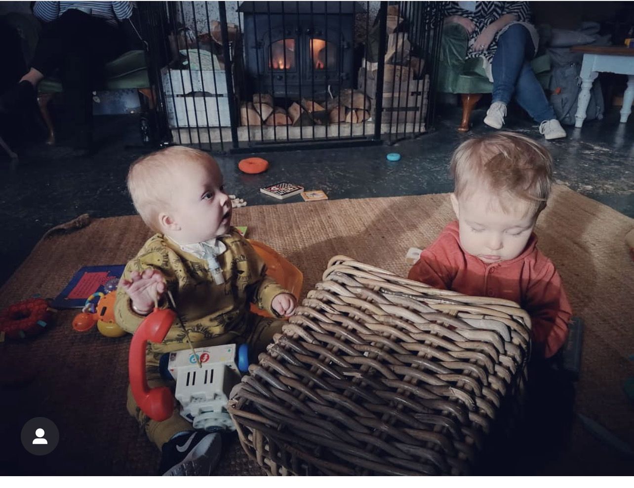 Two young boys playing in a family living room. There is a fireplace with a guard around it behind them and a seagrass rug. The little boy on the left has on a mustard yellow jumper and has a tracheostomy tube. He is holding a plastic toy phone. The other little boy is looking at a toy on his lap and is wearing a red jumper. Both boys have fair brownish hair.