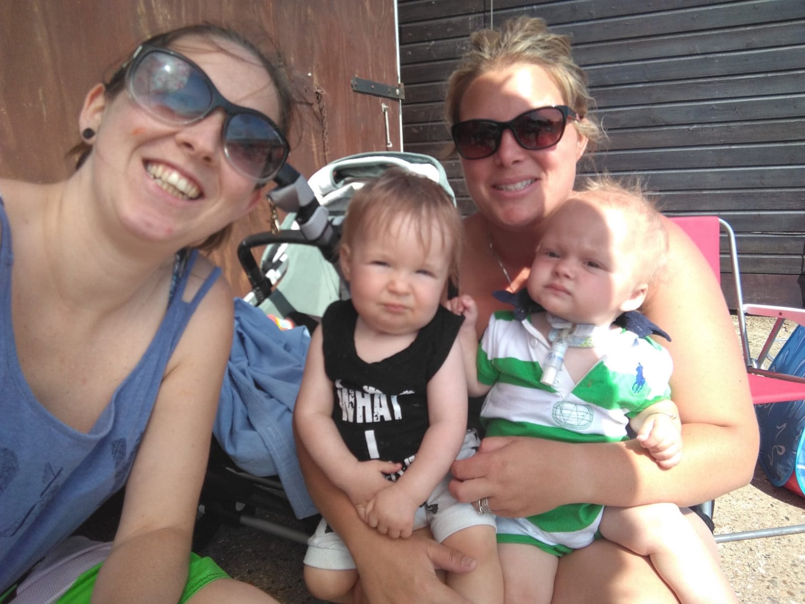 Two mums and their very young children. The mum on the left has on a light blue vest top and dark glasses. She is doing a big smile for the camera. The little boy in the middle has fair brown hair and a black sleeveless t shirt. The other little boy is sitting on his mum's lap. He is wearing a green and white striped t shirt and has a tracheostomy tube. His mum has dark glasses on. It looks like they are at the beach