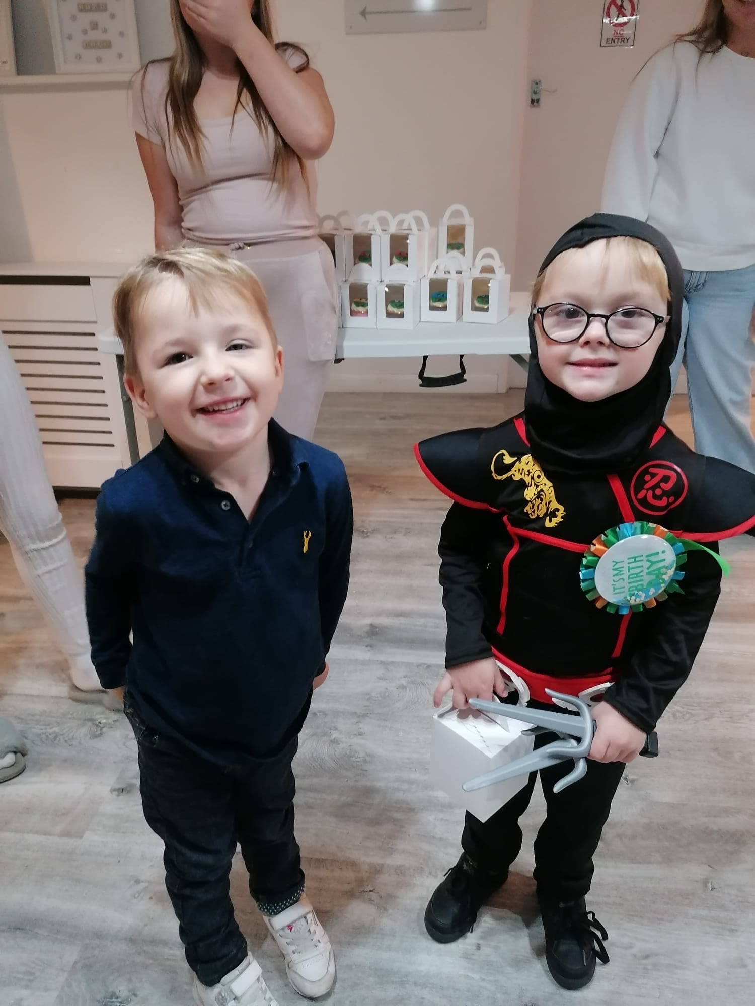 Two young boys standing together. The boy on the left has on a navy shirt and trousers and white trainers. He has blond hair and a side parting. He is smiling. the other boy has on a ninja fancy dress outfit. He has black rimmed glasses.