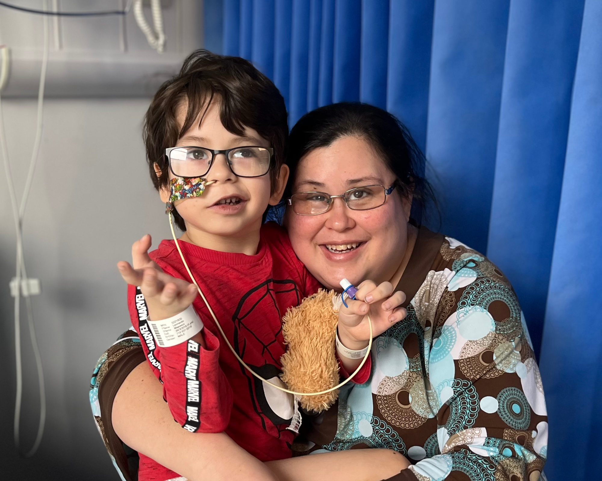 A little boy with dark hair and dark rimmed glasses, wearing a spider man top. He has a nasal tube. He is with his mum who has dark hair and glasses.