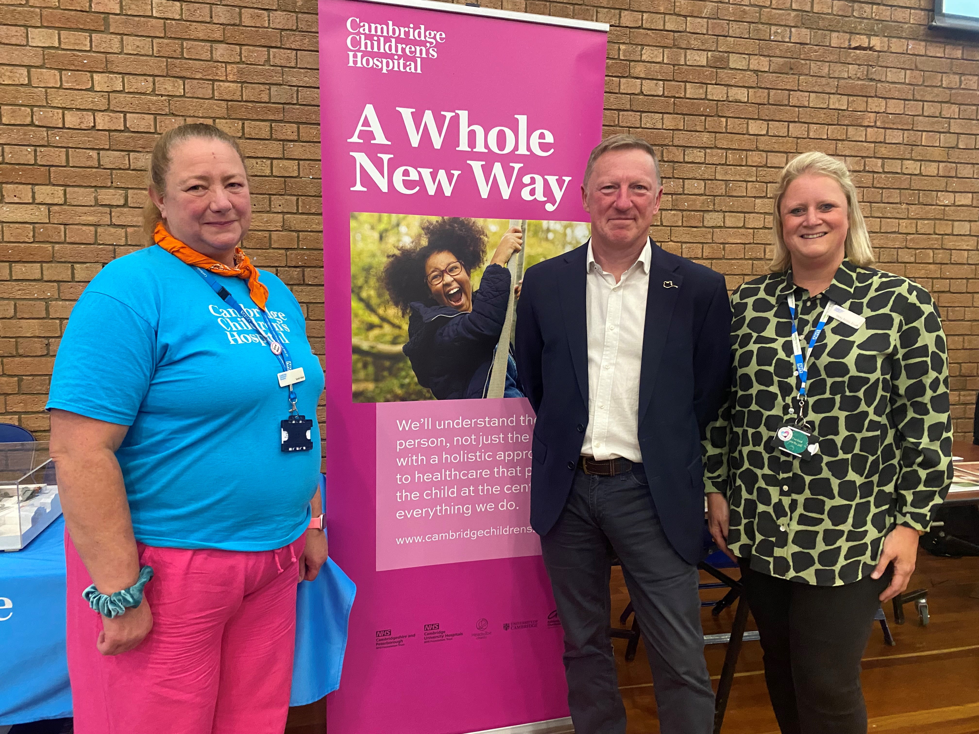 Three people standing with a banner showing a photo of a young girl and the words A Whole New Way. The woman on the left is wearing a bright blue t shirt and pink trousers, with her hair tied up in a pony tail. The man is wearing a dark suit and cream shirt. He has short brown hair. The woman on the left has blond shoulder length hair and is wearing a yellow and black patterned shirt.