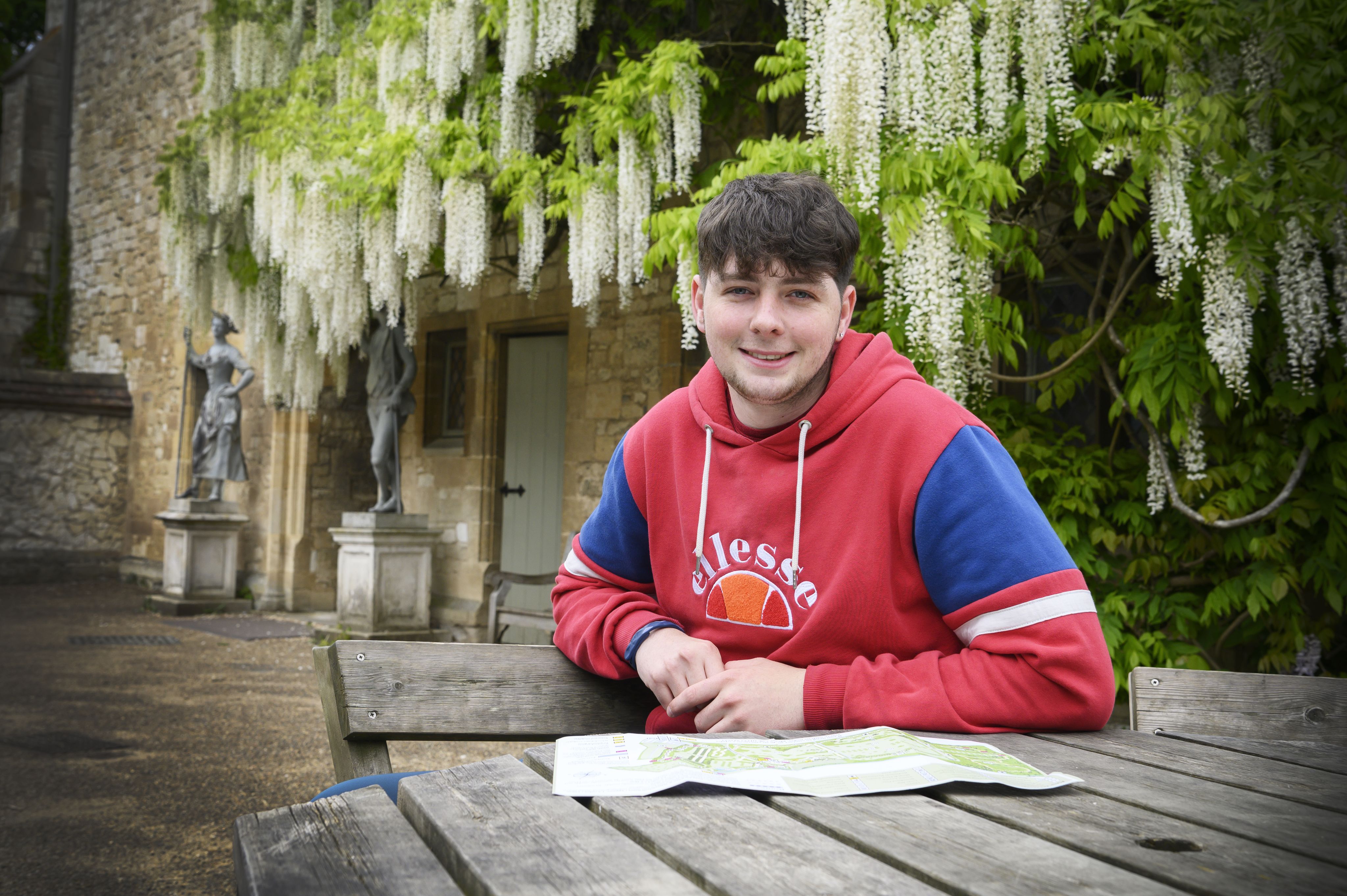 A teenage boy with dark hair and a floppy fringe. He is wearing a red and blue hoodie and has an old building behind him that is covered in wysteria. He is sitting at a wooden table