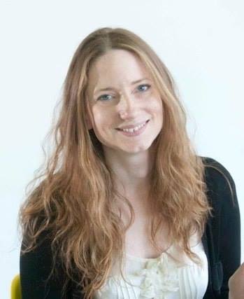 A woman with long reddish brown wavy hair. She is wearing a white top and black cardigan and is smiling