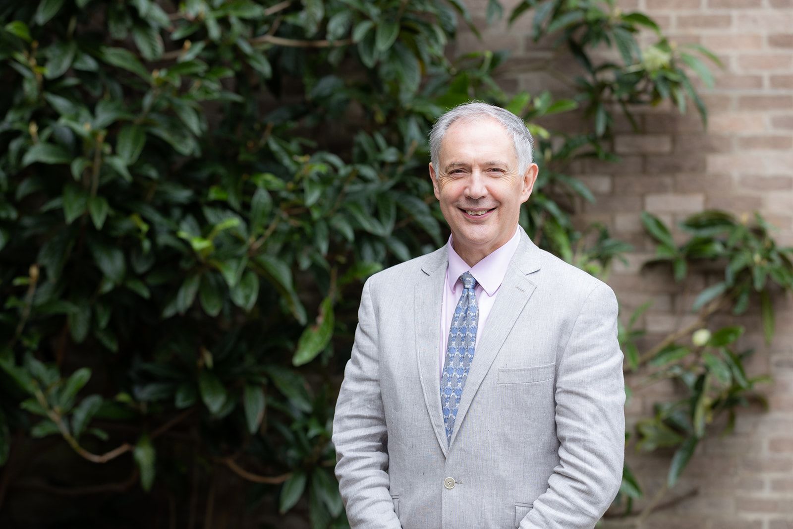 A middle aged man with grey hair, grey suit, pink shirt and pale blue patterned tie. He is smiling and has a brick wall and foliage behind him
