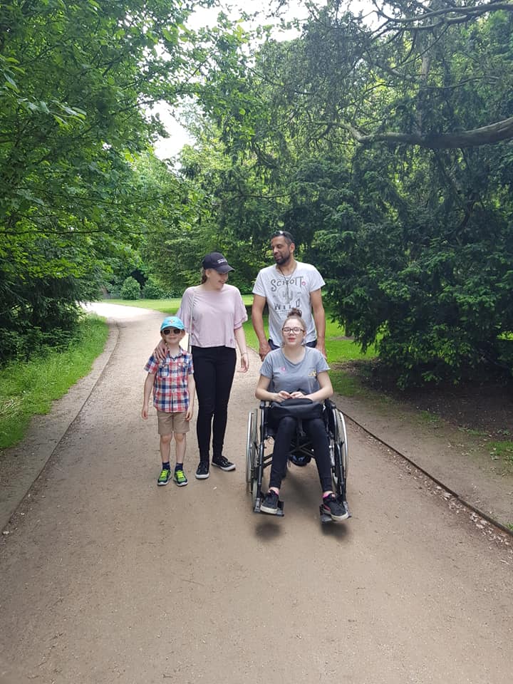Olina in a wheelchair, being pushed by her dad, with her older sister and younger brother. They are on a path, with trees and bushes around them