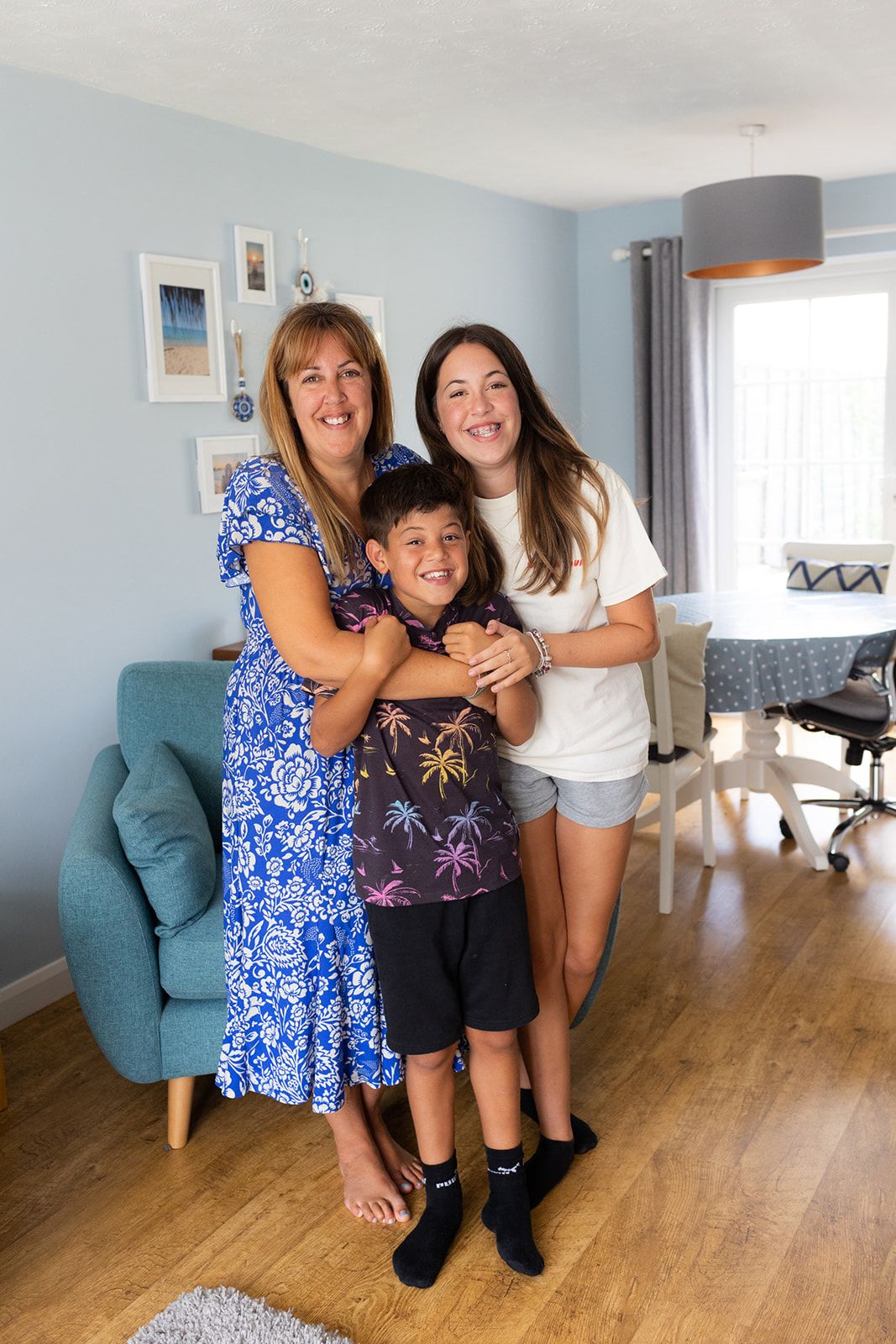 A woman in a long blue flowery summer dress and bare feet. She has long golden brown hair. She is hugging a young boy who has a black patterned t shirt and black shorts. He has short very dark hair. A teenage girl is also in the picture. She has long brown hair, a white t shirt and denim shorts. They are in their living room at home, which has pale blue walls and a wooden floor