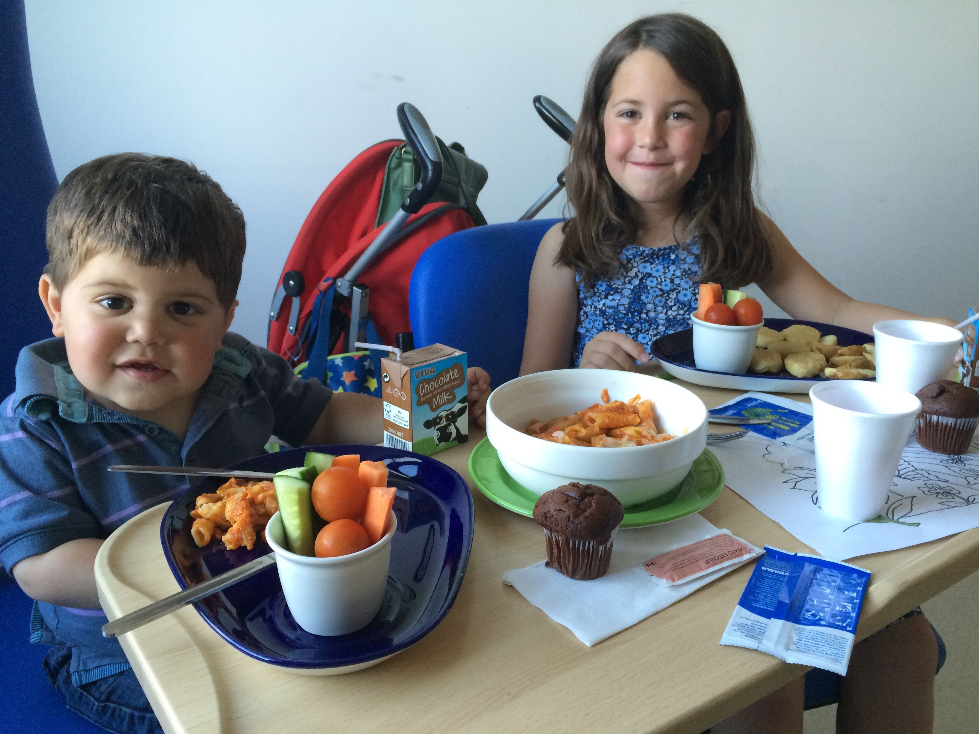 Two children eating their lunch in hospital. The little boy is a toddler, with short brown hair and a blue checked shirt. His sister has long brown hair and a flowery blue top. They are eating penne pasta in tomato sauce, with raw carrots, tomatoes and cucumber. The boy has a chocolate milk carton