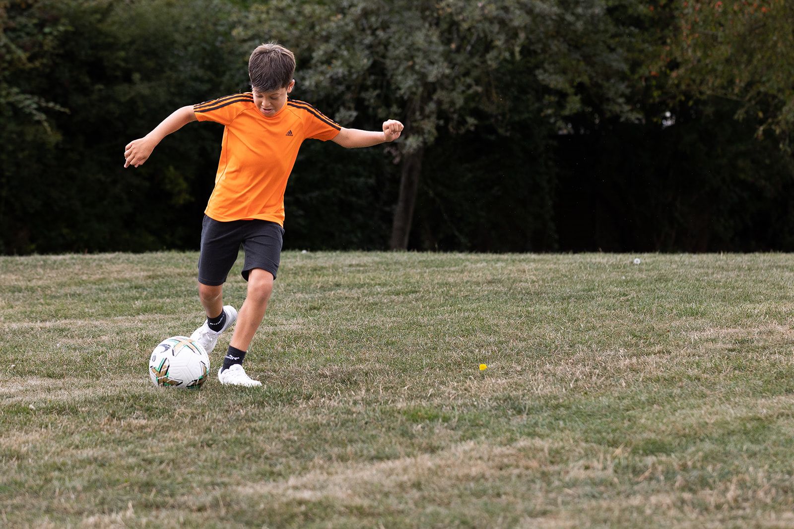 A boy in an orange football top and black shorts playing football in the park