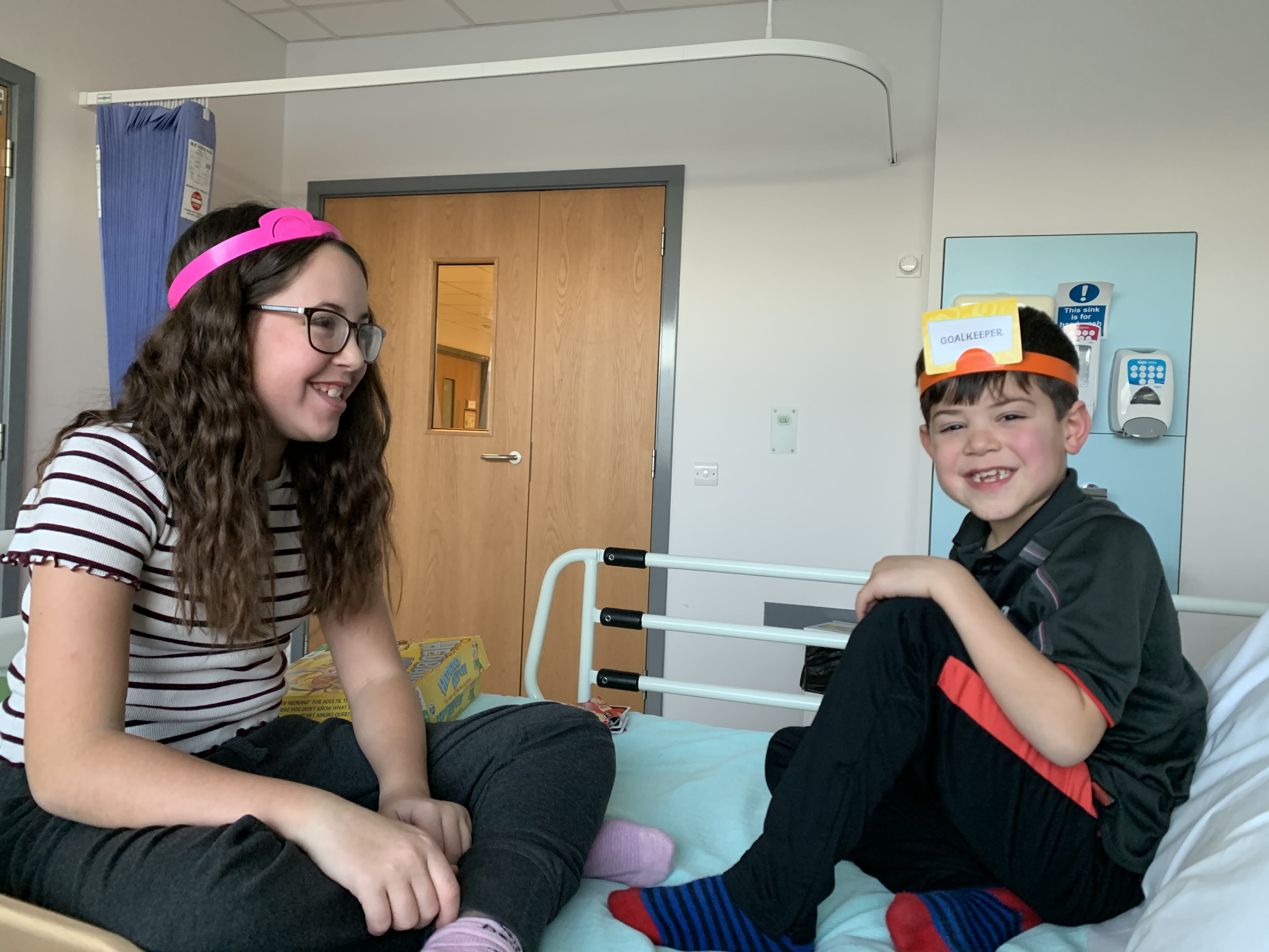 A girl with long brown hair and glasses sitting cross legged on a hospital bed with her brother who has short brown hair and a black football top. They are playing a game where they have to wear headbands with words on, which they have to guess what they are