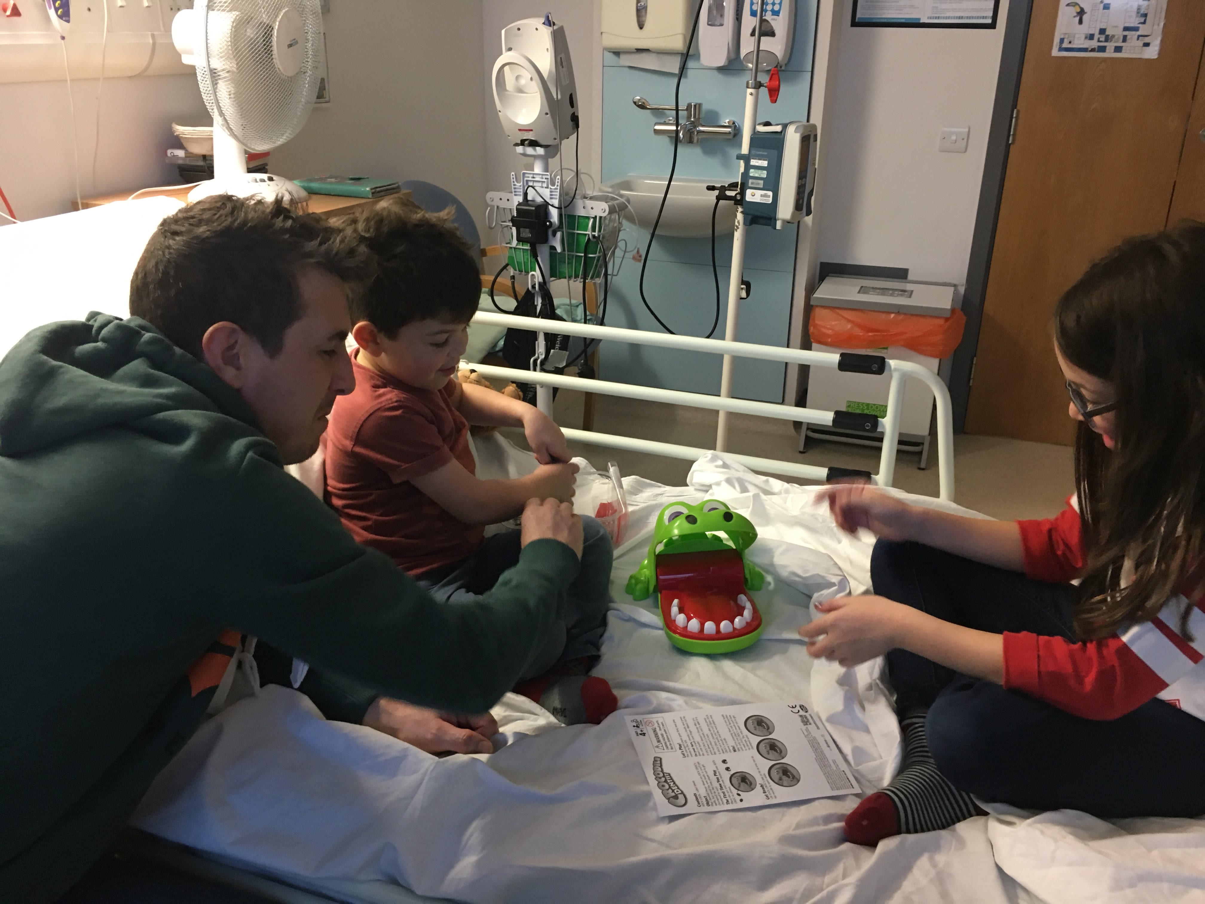 A man playing a game with his children on a hospital bed. The game involves a hungry hippo that they are feeding treats to.