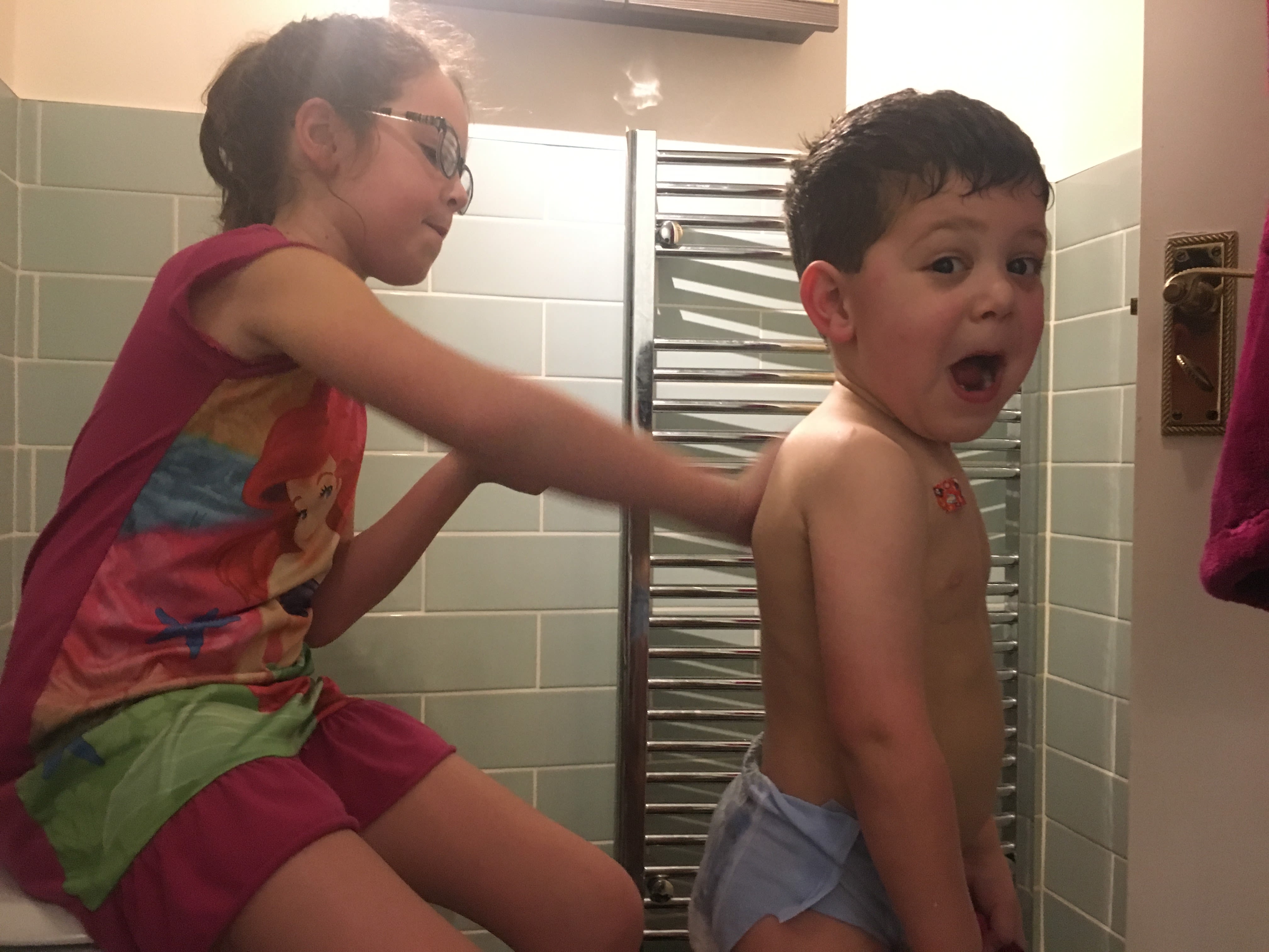 A young girl with brown hair and a pink top is rubbing cream on her little brother's back. He is looking at the camera and pulling a face, as if the cream is cold