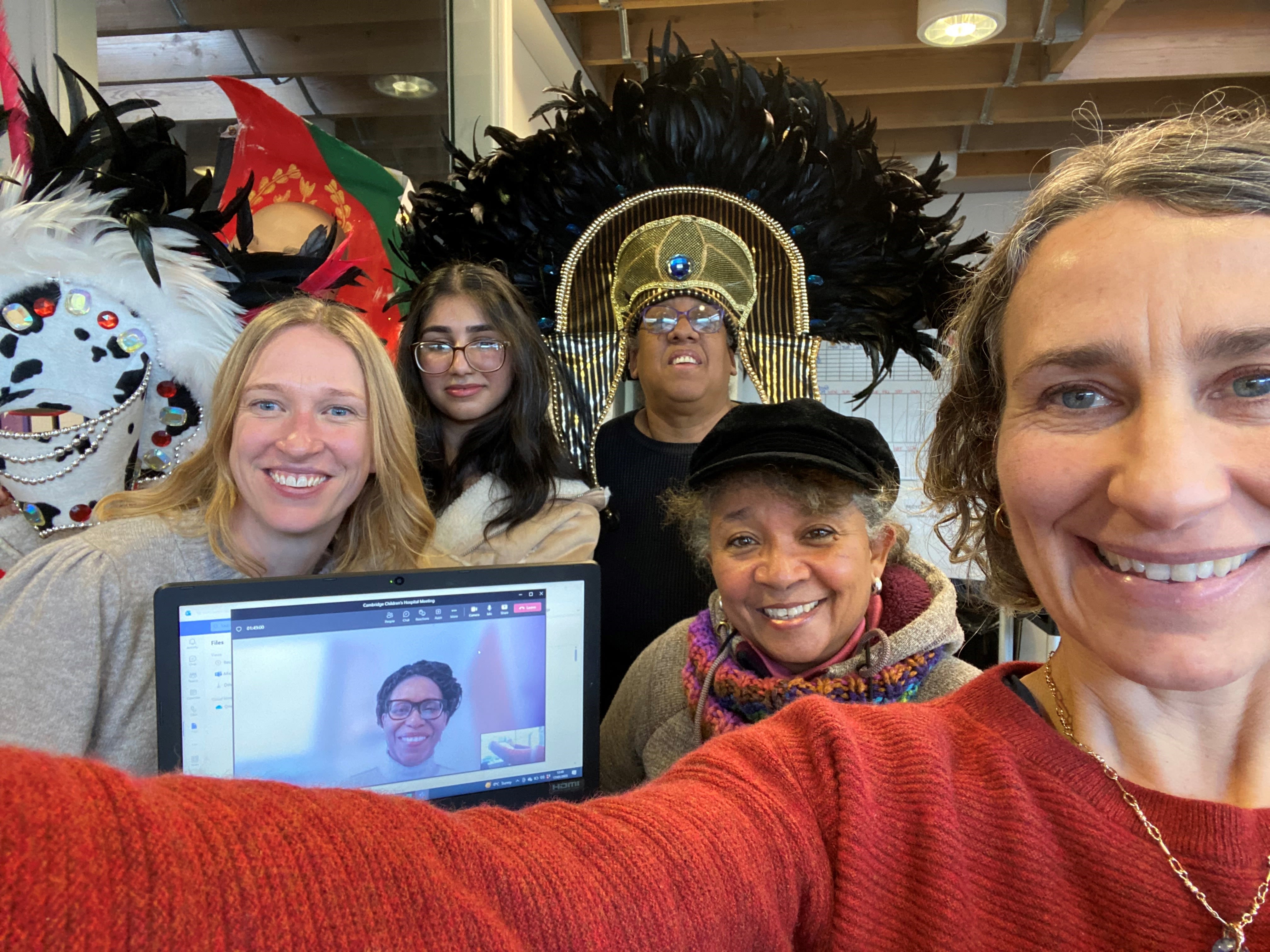 A group of people smiling. One person is on a laptop. A lady is wearing a carnival headdress