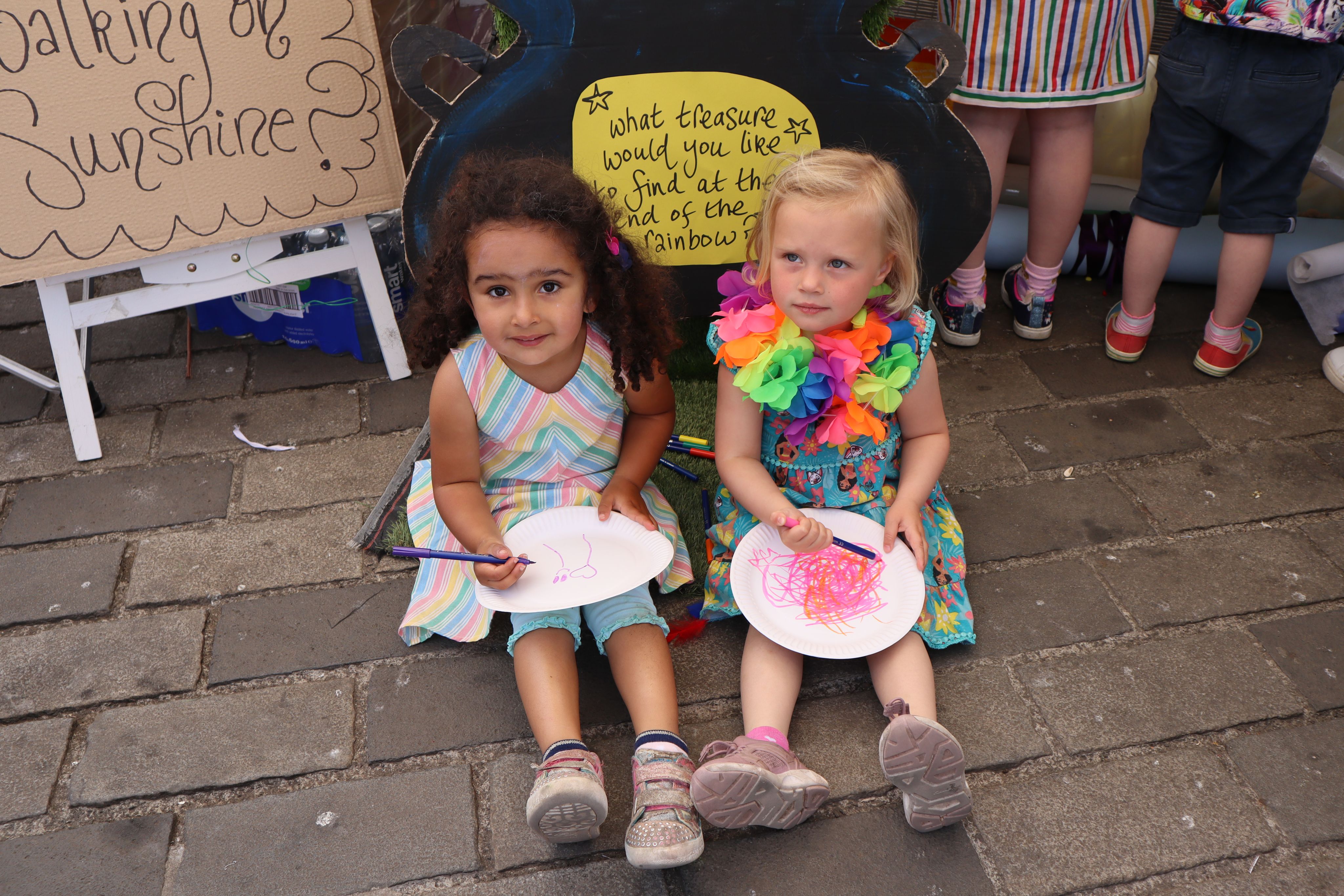 Two little girls colouring on paper plates and smiling at the camera. they are both wearing colourful dresses