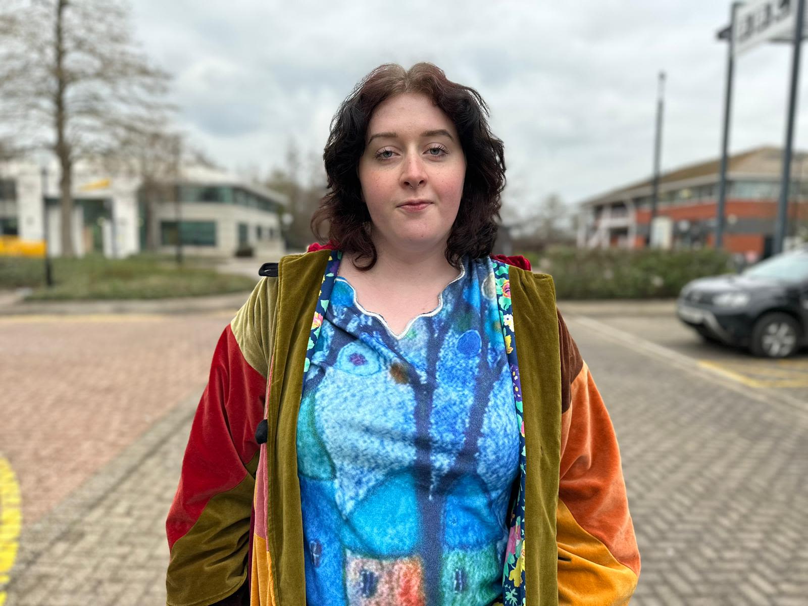 A girl wearing a blue top and orange jacket looking at the camera. She has shoulder length brown hair