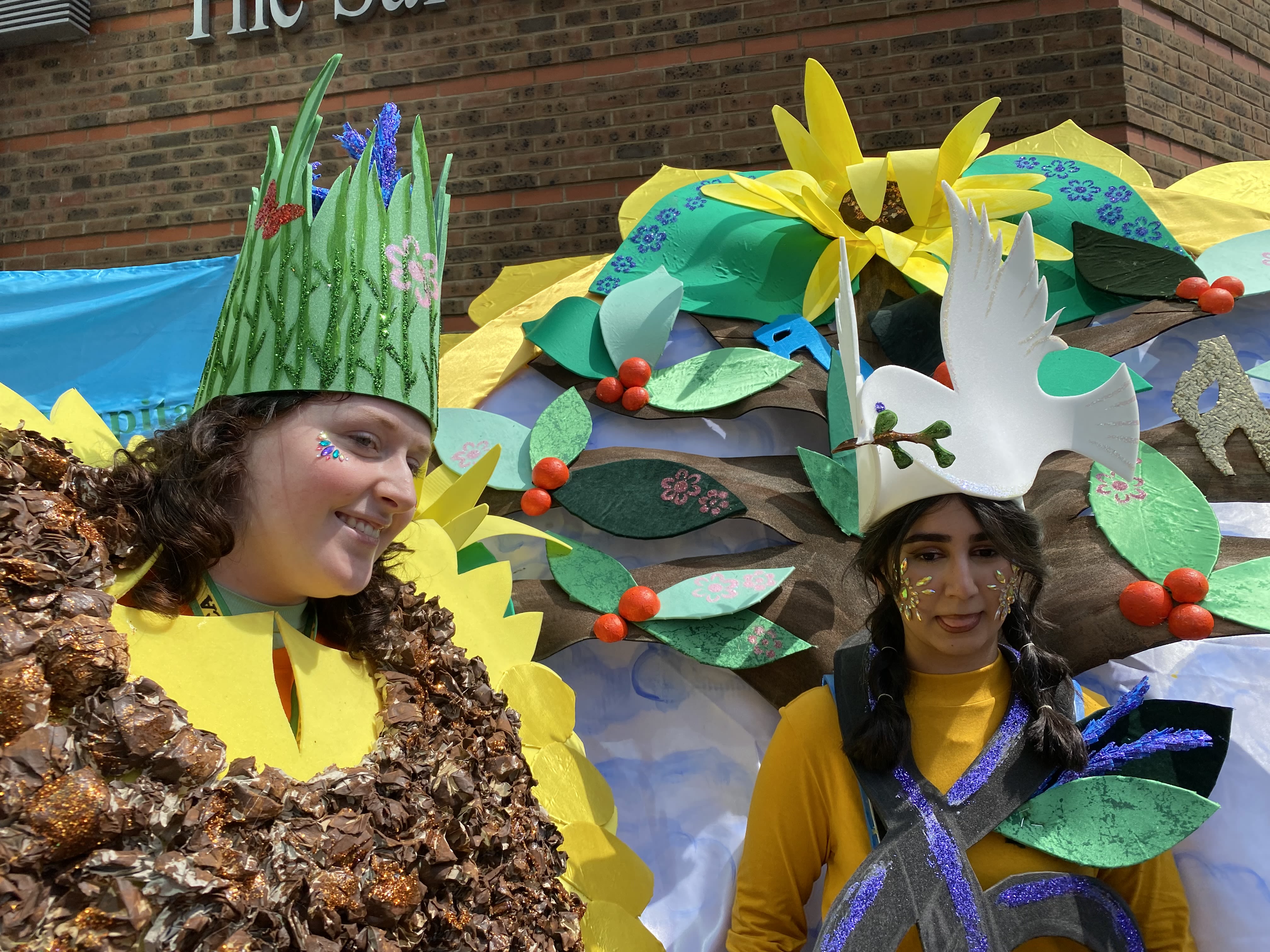 Two teenagrs wearing carnival costumes. One is a sunflower with grass and lavender crown on her head. The other has a dove headdress