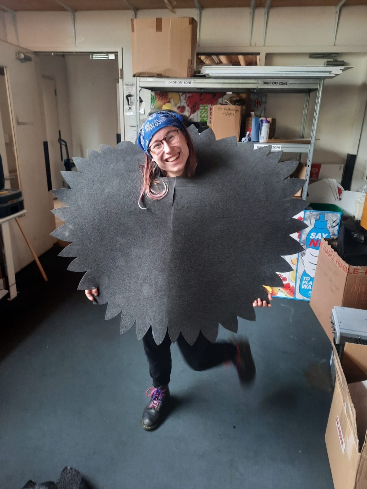 A lady smiling at the camera, wearing a large piece of grey foam shaped like a sunflower
